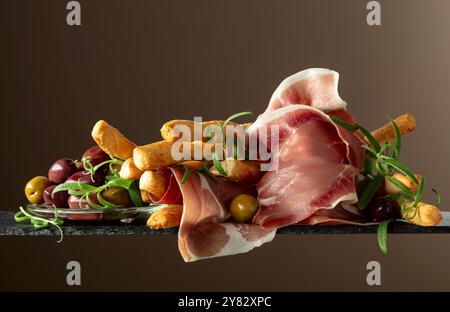 Prosciutto con bastoncini di pane, olive e rosmarino. Tradizionali spuntini italiani su sfondo marrone. Foto Stock