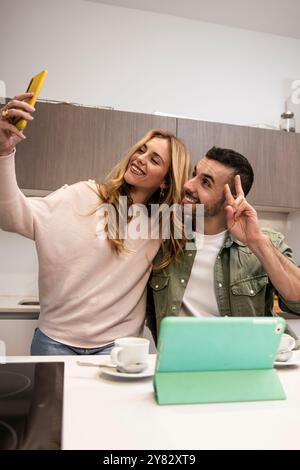 Giovane coppia adulta che usa scattare un selfie da casa in una cucina moderna. Uomo e donna felici e gioiosi in una videochiamata che beve una tazza di caffè. Foto Stock