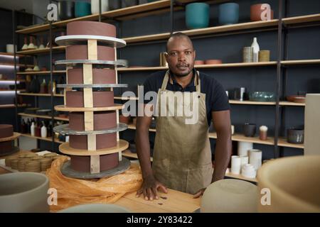 Ritratto di un vasaio africano che indossa un grembiule appoggiato su un banco da lavoro da scaffali pieni di vasi di argilla in uno studio di ceramica Foto Stock