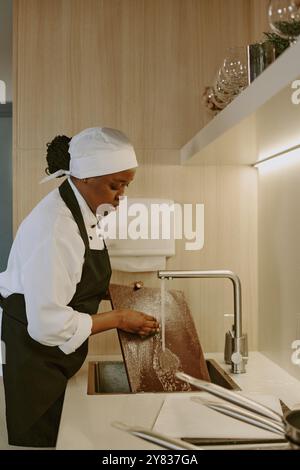 Cuoco afroamericano in uniforme bianca con grembiule nero lavatrice tagliere nella moderna cucina leggera del ristorante Foto Stock