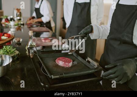 Primo piano medio di mani maschili irriconoscibili con guanti neri che usano pinze per capovolgere la bistecca di manzo Foto Stock