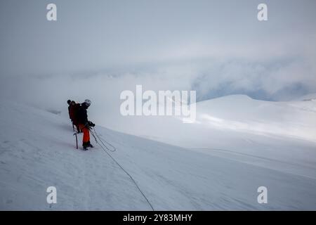 Solitario scalatore braves vasta distesa innevata: Alpinista legato alla sicurezza, oscurato dall'immensa e bianca natura selvaggia di Kazbek. Foto Stock