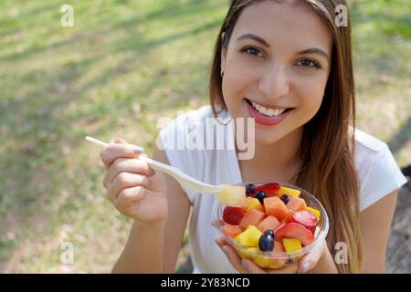 Bella giovane donna che mangia un'insalata di frutta fresca con frutta diversa e colorata nel parco cittadino. Guardando la telecamera. Foto Stock