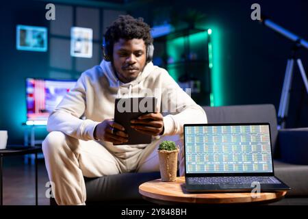 L'uomo ascolta la musica e legge dal tablet, aspettando che i dati finanziari del mercato azionario sullo schermo del notebook si aggiornino. Uomo d'affari a casa accanto a un notebook che mostra i grafici degli investimenti commerciali Foto Stock