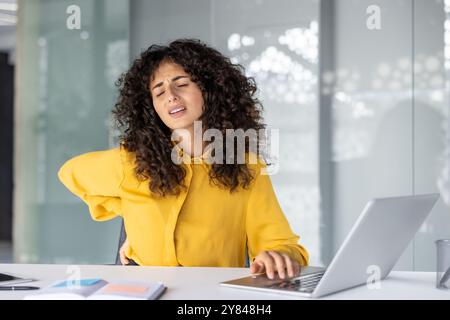 Donna d'affari ispanica che sente disagio per mal di schiena seduta alla scrivania in ufficio. La donna mostra segni di tensione, lavora su computer portatile e telefono. Riflette le sfide dell'ergonomia sul luogo di lavoro Foto Stock