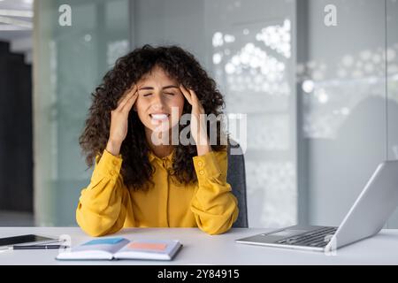 Donna d'affari con gli occhi chiusi che tiene la testa in mano, sente stress e mal di testa alla scrivania. Sopraffatta dal carico di lavoro, si trova in ufficio con un notebook, un telefono e un notebook aperto. Foto Stock