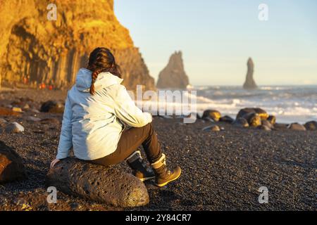 Escursionista donna seduta sulla spiaggia di sabbia nera di Reynisfjara che guarda il mare. Islanda Foto Stock