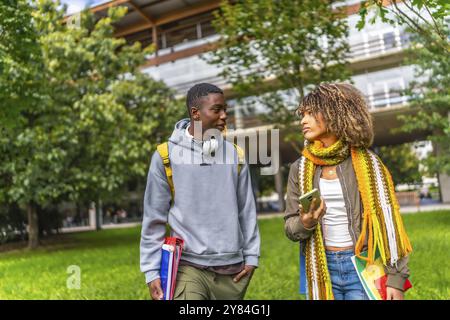 Vista frontale di due studenti multietnici che chiacchierano e usavano i dispositivi mobili che uscivano dall'università Foto Stock