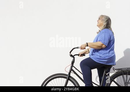 Donna matura sorridente in sovrappeso in sella a una bicicletta in stile retrò isolata su sfondo bianco e spazio di copia, concetto di esercizio negli anziani Foto Stock