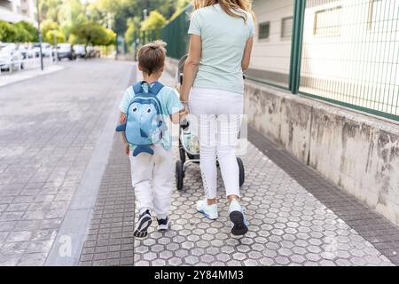 Vista posteriore di una madre e un figlio che trasportano una carrozza per bambini che camminano insieme fino a scuola Foto Stock