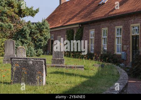 Cimitero con vecchie lapidi di fronte a edifici in mattoni rossi con tetti rossi e vegetazione verde, krummhoern, frisia orientale, germania Foto Stock