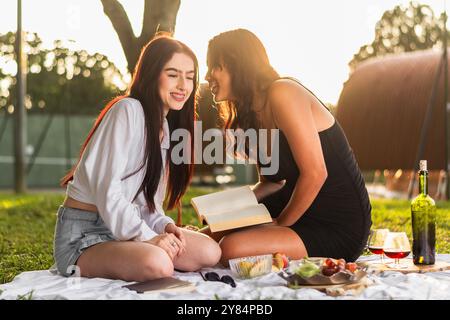 due amici latini sorridenti e felici mentre si godono il picnic, parlano e raccontano segreti nelle orecchie dell'altro, tramonta sullo sfondo. Foto Stock