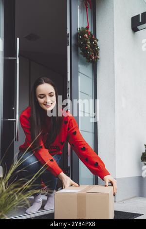 Giovane donna allegra eccitata per un pacco che ha appena ricevuto. Donna che riceve il suo regalo di natale per posta. Aprendo il regalo di Natale a casa Foto Stock