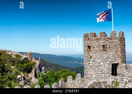 Castello moresco merlature in pietra e bandiera portoghese Sintra // SINTRA, Portogallo - la storica torre di guardia e merlature in pietra del castello moresco (Castelo dos Mouros) si trovano in cima alla collina di Sintra, con la bandiera portoghese che sventola sopra. Questa fortificazione medievale fu costruita durante l'VIII e il IX secolo dai Mori che occuparono la penisola iberica. La posizione strategica del castello offriva vedute dominanti della regione circostante e serviva come avamposto militare. In seguito alla riconquista cristiana, la fortezza subì diversi lavori di ristrutturazione, tra cui una significativa r Foto Stock