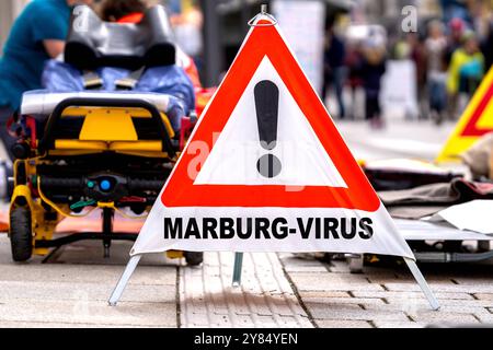 Germania - 3 ottobre 2024: Segnale di avvertimento: Virus di Marburg, di fronte ai paramedici di emergenza in una strada. Immagine simbolica del virus pericoloso che si diffonde e infetta gli altri. FOTOMONTAGGIO *** Warnschild: Marburg-virus, vor Notfallsanitätern auf einer Straße. Symbolbild Notfall und Ansteckung des gefährlichen virus. FOTOMONTAGE Foto Stock