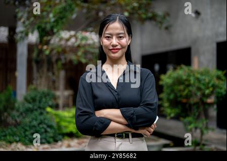 Ritratto di una donna d'affari asiatica sicura e attraente in un abito elegante, in piedi con le braccia incrociate in giardino e sorridente alla macchina fotografica. Foto Stock