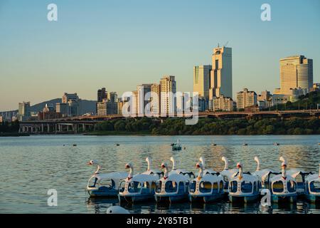Questa foto cattura una tranquilla mattinata in un parco, con barche a forma di cigno allineate sul fiume calmo e sullo sfondo lo skyline della città. Foto Stock