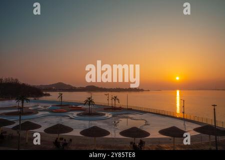 Un tranquillo tramonto dorato getta riflessi sul mare calmo, con palme e ombrelloni che creano un'atmosfera tranquilla per il relax. Foto Stock