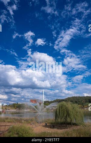 Un bellissimo paesaggio caratterizzato da una fontana e da alberi lussureggianti sotto un cielo blu luminoso pieno di nuvole, con il tranquillo lago che riflette il paesaggio. Foto Stock