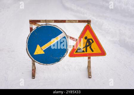 Un segnale di avvertimento per i lavori in corso e un segnale di deviazione sono posizionati su una strada innevata, che avvisa i conducenti dei lavori stradali in programma e li guida ad alternarsi Foto Stock