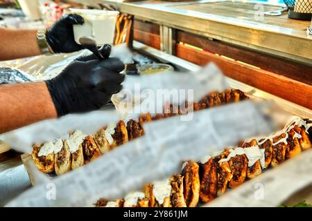 Uno chef sta preparando un lungo sandwich con vari ingredienti in un evento all'aperto, circondato da erbe fresche e prodotti alimentari. Foto Stock