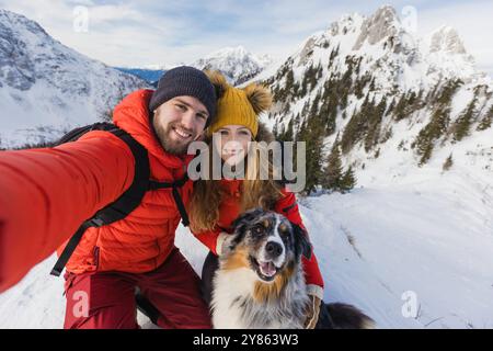 La coppia di escursionisti con un cane scatta un selfie da un paesaggio innevato nelle montagne invernali Foto Stock