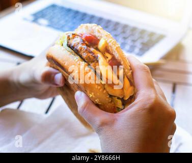 Mani che reggono un gustoso hamburger con patatine fritte e formaggio fuso con un notebook sfocato sullo sfondo Foto Stock