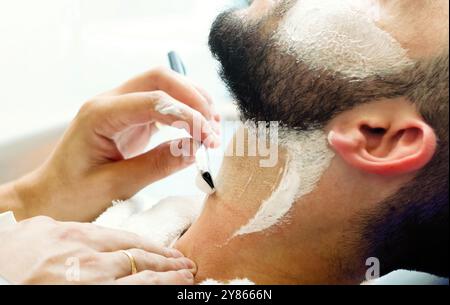 Barber rasa con cura la barba di un cliente con un rasoio liscio, applicando la crema da barba per una rasatura precisa e liscia Foto Stock