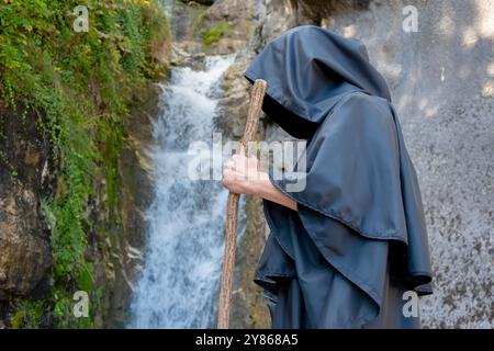 Un monaco in abito nero con un personale passa davanti a una cascata di montagna. Foto Stock