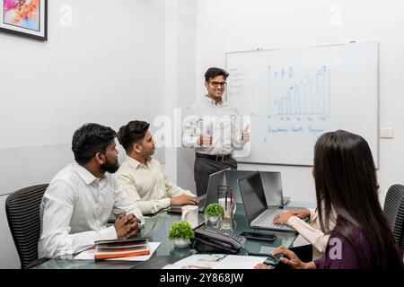 Questa immagine rappresenta una dinamica riunione del team di lavoro indiano in un ambiente elegante e moderno. La figura centrale, un giovane capo indiano con expre Foto Stock