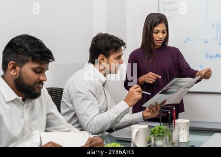 Colleghi diversi che lavorano con documenti finanziari, statistiche di progetto in sala riunioni insieme, team leader indiano-americano che tiene briefing, dipendente. Foto Stock