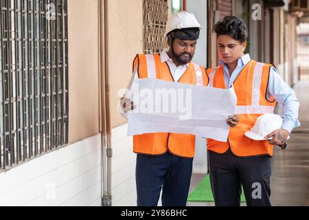 Lavoro di squadra mirato in cui due ingegneri di progetto e architetti focalizzati in un cantiere edile, esaminando diligentemente un progetto o una planimetria indossati entrambi Foto Stock