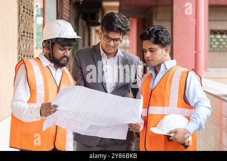 Un architetto, un ingegnere civile e un interior designer che esaminano da vicino una vasta serie di progetti edilizi in un vivace cantiere e ne discutono. Foto Stock