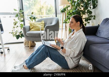 Una giovane donna siede sul pavimento, vestita in modo informale, impegnata in lavori a distanza in un ambiente accogliente e luminoso. Foto Stock