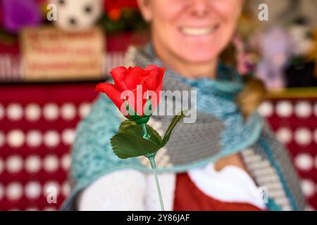 Monaco di Baviera, Germania - 23 settembre 2024: Una donna, operatore di una galleria di tiro all'Oktoberfest, consegna in premio una rosa rossa artificiale. Indossa il tradizionale costume bavarese, mentre lo stand può essere visto sullo sfondo *** Eine Frau, Betreiberin einer Schießbude auf dem Oktoberfest, überreicht eine rote Kunstrose als Gewinn. SIE trägt bayerische Tracht, während im Hintergrund der Stand zu sehen ist Foto Stock