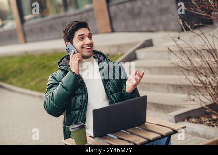 Foto di un uomo positivo che siede sulla terrazza del caffè all'aperto e parla al telefono mentre si gode il fine settimana all'aperto Foto Stock