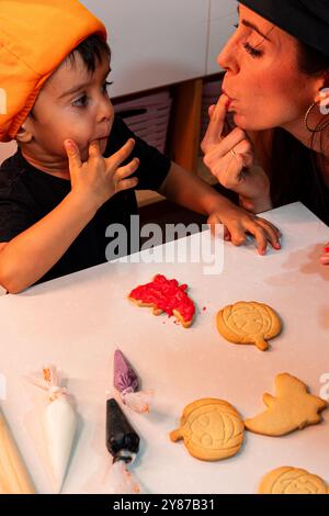 Una donna e un bambino stanno facendo i biscotti di Halloween. La donna sta mettendo la ciliegina su un biscotto mentre il bambino si lecca le dita Foto Stock