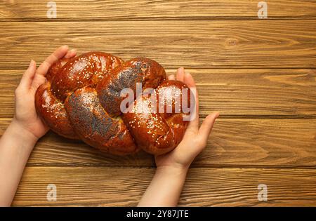 Mani femminili con pane Challah appena sfornato ricoperto di papavero e semi di sesamo, vista dall'alto su uno sfondo rustico di legno, tradizionale festivo ebraico Foto Stock