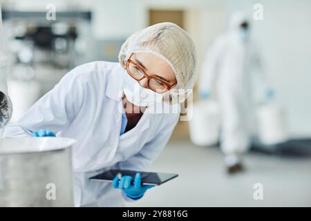 Supervisore di produzione femminile in maschera protettiva e occhiali che supervisiona le apparecchiature controllando il processo di produzione e documentando i risultati in tablet digitali presso l'impianto farmaceutico, spazio di copia Foto Stock