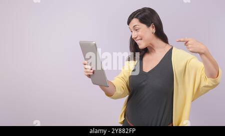 Una donna con un'espressione allegra tiene in mano un tablet e lo indica. Indossa un maglione giallo sopra una camicia nera e l'immagine viene scattata Foto Stock