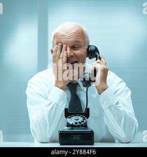 Un uomo d'affari più anziano viene colto alla sprovvista di una conversazione che sta avendo su un telefono fisso vintage, coprendo il suo viso con la mano mentre sorride Foto Stock