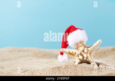 Stelle marine nel cappello di Babbo Natale sulla spiaggia di sabbia su sfondo blu. Un'idea creativa per le vacanze tropicali di Natale. Foto Stock