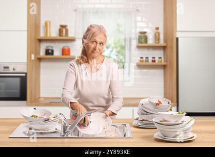 Donna stanca che lavava i piatti in un lavandino su un bancone di legno in una cucina Foto Stock