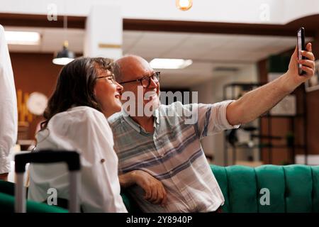Vecchio anziano che tiene il cellulare per un selfie con la moglie nell'esclusiva hall dell'hotel. Coppia anziana in pensione con valigia, seduta nell'area salotto con dispositivo mobile in mano per una videochiamata. Foto Stock