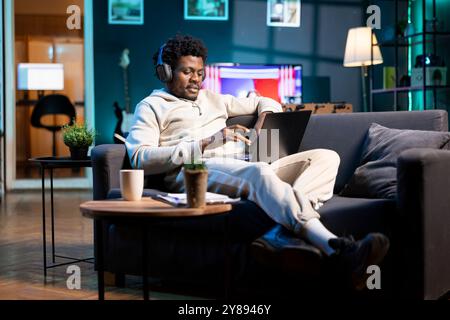 Lavoratore remoto felice che ascolta la musica con le cuffie mentre lavora da casa. Libero professionista sorridente e rilassato che si diverte, scrivendo al computer nell'elegante ufficio personale dell'appartamento Foto Stock