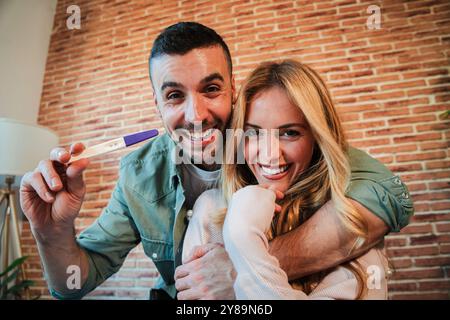 Coppia eccitata che celebra il test di gravidanza positivo, che abbraccia e sorride insieme a casa, condivide la gioiosa notizia dell'aspettarsi un bambino, esprimendo felicità e attesa per il loro nuovo viaggio in famiglia. Foto di alta qualità Foto Stock