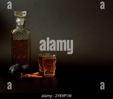 Liquore di prugne con cannella e anice su sfondo nero, alcol forte fatto in casa in vetro di cristallo, frutta matura e spezie. Primo piano. Foto Stock