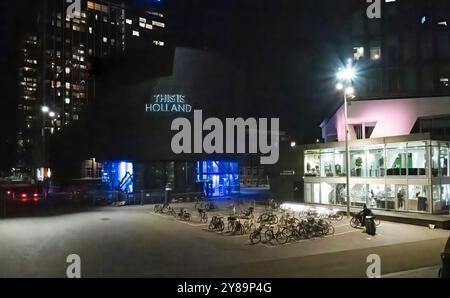 Amsterdam, Paesi Bassi - 15 settembre 2024: Desolato parcheggio per biciclette di notte illuminato da un edificio con facciata in vetro, accanto a questa attrazione Holland. Foto Stock