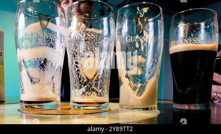 Quattro bicchieri di birra Guinness Stout; tre bicchieri vuoti e un bicchiere con schiuma cremosa e birra scura; logo dell'arpa Brian Boru. Foto Stock