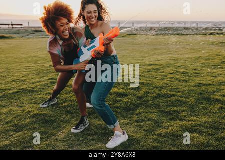 Amici gioiosi che giocano con le pistole ad acqua nel parco cittadino al tramonto, catturando l'essenza del divertimento, della risate e dell'amicizia in un ambiente all'aperto spensierato Foto Stock